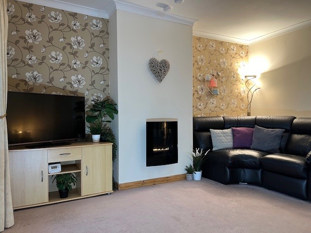 Photo of Easterfield Road front living room featuring a modern built in fireplace, a sofa, a TV Unit and TV. The walls are papered with a tasteful floral pattern and plain around the chimney breast